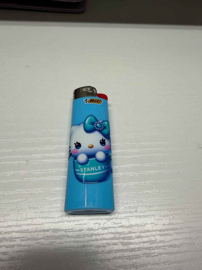 以下が含まれることがあります： ハローキティのデザインが施された青いBICライター。ライターには赤い点火ボタンと、名前「Stanley」がプリントされた白と青のハローキティのグラフィックがあります。