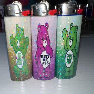 Puede incluir: Tres encendedores cubiertos de purpurina con dise&ntilde;os estilo Ositos Cari&ntilde;osos. Un oso verde dice "Eat Shit", un oso morado dice "Kiss My Ass" y un oso verde dice "Fuckin". Cada encendedor tiene una parte superior plateada y un bot&oacute;n rojo.