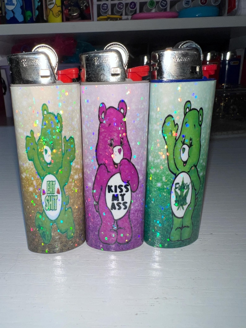 Puede incluir: Tres encendedores cubiertos de purpurina con dise&ntilde;os estilo Ositos Cari&ntilde;osos. Un oso verde dice "Eat Shit", un oso morado dice "Kiss My Ass" y un oso verde dice "Fuckin". Cada encendedor tiene una parte superior plateada y un bot&oacute;n rojo.