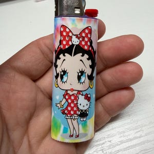 May include: A cylindrical lighter featuring a colorful tie-dye background with a graphic of Betty Boop wearing a red dress with white patterns and a matching bow, holding a small Hello Kitty figure. The lighter has a metallic top and red ignition button.
