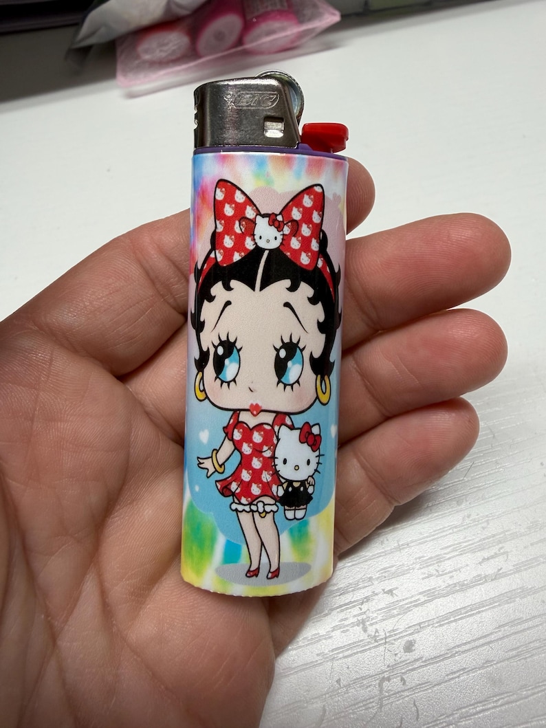 May include: A cylindrical lighter featuring a colorful tie-dye background with a graphic of Betty Boop wearing a red dress with white patterns and a matching bow, holding a small Hello Kitty figure. The lighter has a metallic top and red ignition button.