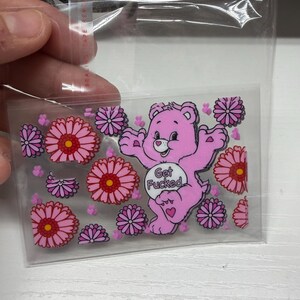 Op de afbeelding: Een doorzichtige plastic hoes met een rechthoekige sticker van een roze cartoonbeer met een wit buikembleem met de tekst Get Fucked, omringd door talloze roze en paarse bloemen en kleine roze hartjes als accenten.