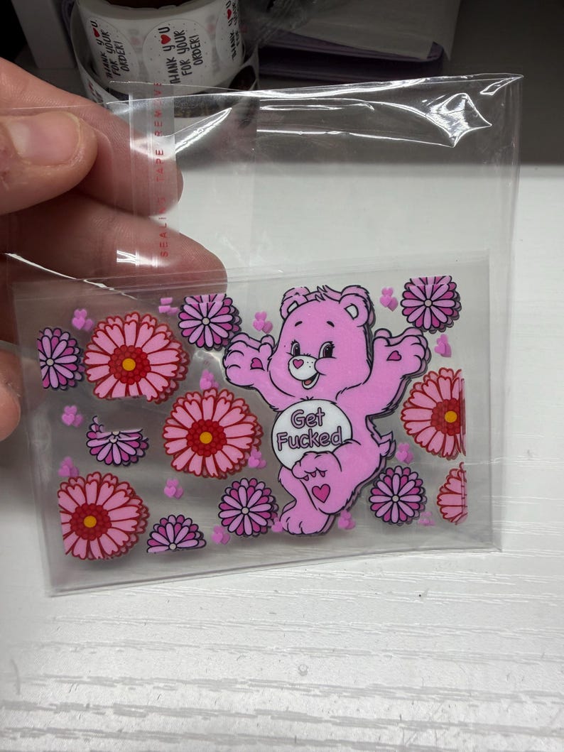 Op de afbeelding: Een doorzichtige plastic hoes met een rechthoekige sticker van een roze cartoonbeer met een wit buikembleem met de tekst Get Fucked, omringd door talloze roze en paarse bloemen en kleine roze hartjes als accenten.