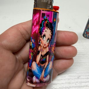 May include: A Bic lighter featuring a colorful graphic of a cartoon character with rainbow-streaked hair, wearing a black off-the-shoulder top and blue jeans. The text Sassy & Classy is printed in pink neon-style script above the character.