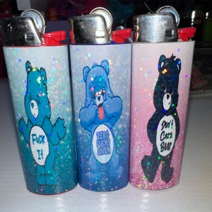 Puede incluir: Tres encendedores cubiertos de purpurina con dise&ntilde;os de osos de dibujos animados. Uno es azul con el texto "Fuck It", otro es azul con "Zero Fucks Given", y el &uacute;ltimo es negro con "Don't Care Bear".