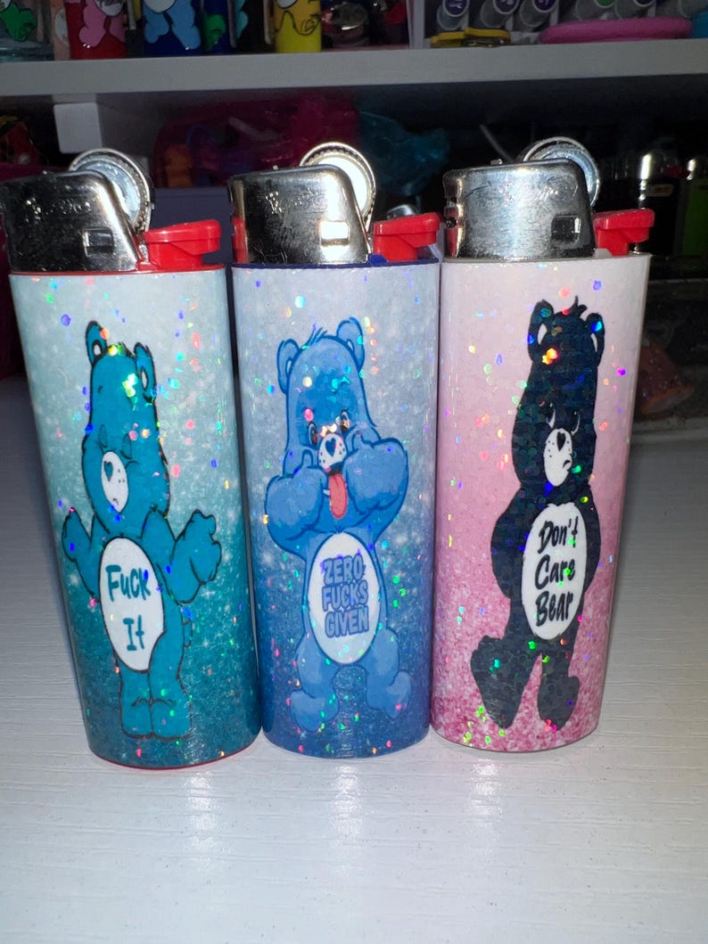 Puede incluir: Tres encendedores cubiertos de purpurina con dise&ntilde;os de osos de dibujos animados. Uno es azul con el texto "Fuck It", otro es azul con "Zero Fucks Given", y el &uacute;ltimo es negro con "Don't Care Bear".