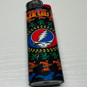 Puede incluir: Un encendedor negro con un bot&oacute;n rojo y una parte superior de metal. El encendedor presenta un dise&ntilde;o colorido con el texto "Grateful Dead" y una calavera con un rayo. El dise&ntilde;o tambi&eacute;n incluye osos bailarines.