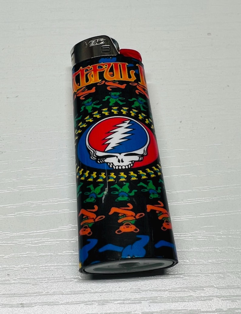 Puede incluir: Un encendedor negro con un bot&oacute;n rojo y una parte superior de metal. El encendedor presenta un dise&ntilde;o colorido con el texto "Grateful Dead" y una calavera con un rayo. El dise&ntilde;o tambi&eacute;n incluye osos bailarines.