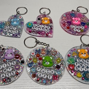 May include: Six glittery keychains with Care Bear-like characters and phrases like "Hot Mess," "Smile Bitch," and "Fuck It." Each keychain has a silver keyring and is decorated with colorful beads and glitter.