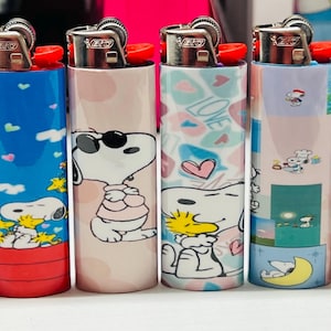 May include: Six decorative lighters featuring Snoopy and Woodstock designs. The lighters have various backgrounds, including pink, blue, and patterns with hearts and the word "LOVE". Each lighter has a red ignition button.