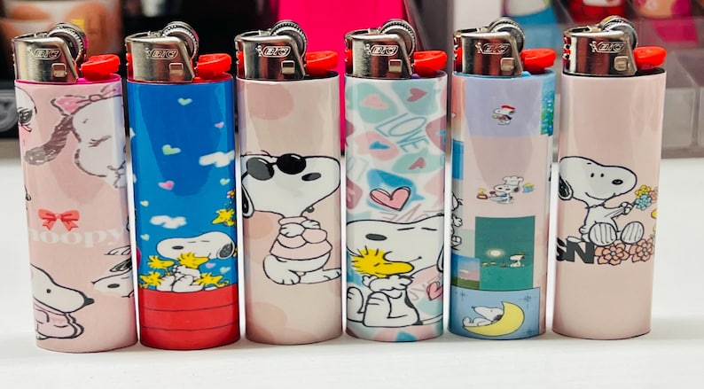 May include: Six decorative lighters featuring Snoopy and Woodstock designs. The lighters have various backgrounds, including pink, blue, and patterns with hearts and the word "LOVE". Each lighter has a red ignition button.