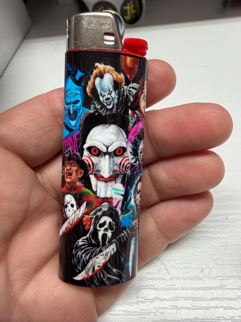 Peut inclure: Un briquet Bic standard dot&eacute; d'un habillage noir orn&eacute; d'un collage de divers personnages de films d'horreur, incluant un masque de marionnette blanc central aux joues en spirale rouge, une silhouette encapuchonn&eacute;e avec un couteau et d'autres figures.