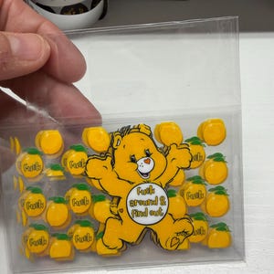Op de afbeelding: Een doorzichtig plastic zakje met een verzameling gele stickers. De stickers tonen een geel beerfiguurtje met de tekst "Fuck around & find out" op de buik, omringd door kleinere gele perzikstickers met het woord "Fuck" erop gedrukt.