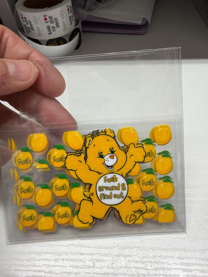 Op de afbeelding: Een doorzichtig plastic zakje met een verzameling gele stickers. De stickers tonen een geel beerfiguurtje met de tekst "Fuck around & find out" op de buik, omringd door kleinere gele perzikstickers met het woord "Fuck" erop gedrukt.