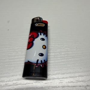 以下が含まれることがあります： ハローキティのデザインが施された、白と黒のBICライター。ライターには赤いボタンと銀色の金属製のトップが付いています。ハローキティのグラフィックには、赤いリボンと黒いひげが含まれています。