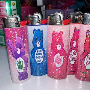 Puede incluir: Varios encendedores cubiertos de purpurina con dise&ntilde;os de osos de dibujos animados. Los encendedores son de varios colores, incluyendo morado, azul y rosa, con textos como "Fuck Around And Find Out".