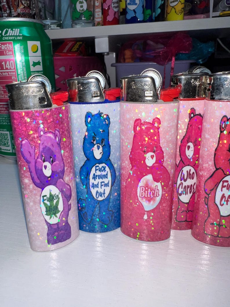 Puede incluir: Varios encendedores cubiertos de purpurina con dise&ntilde;os de osos de dibujos animados. Los encendedores son de varios colores, incluyendo morado, azul y rosa, con textos como "Fuck Around And Find Out".