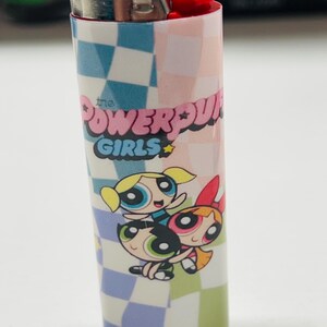 May include: A BIC lighter with a design featuring "The Powerpuff Girls" text and cartoon characters. The lighter has a red button and a silver metal top, with a checkered pattern in blue, pink, and white.