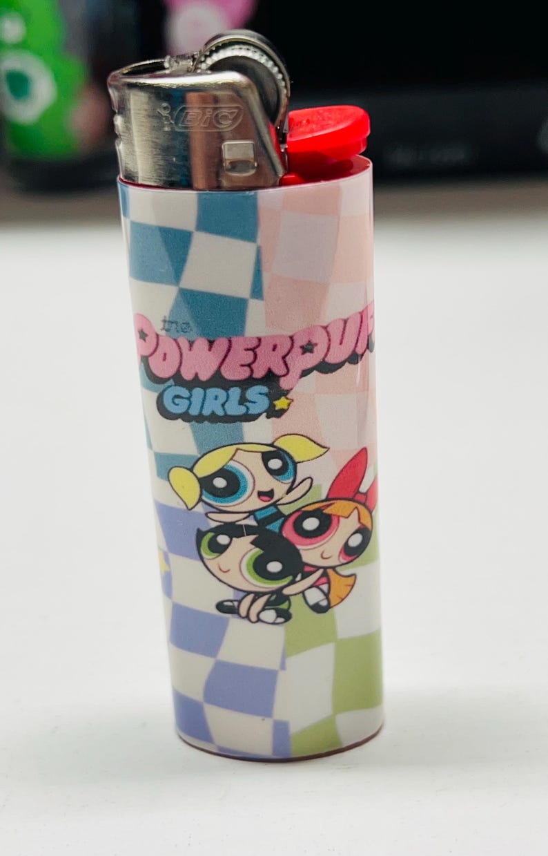 May include: A BIC lighter with a design featuring "The Powerpuff Girls" text and cartoon characters. The lighter has a red button and a silver metal top, with a checkered pattern in blue, pink, and white.