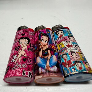 May include: Three rectangular lighters featuring Betty Boop graphics. The left lighter is pink with the word love and Betty Boop. The center lighter shows Betty Boop in blue jeans and a black top. The right lighter features a comic strip design.