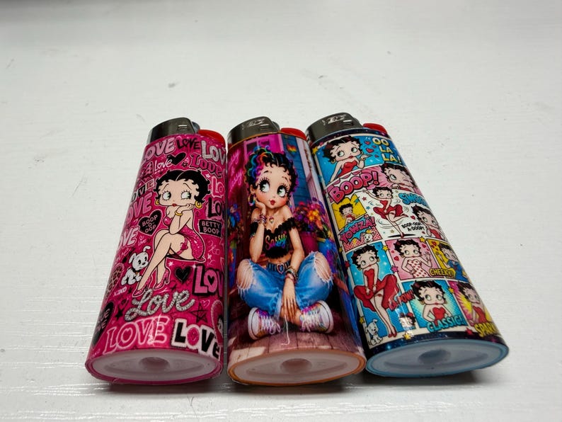 May include: Three rectangular lighters featuring Betty Boop graphics. The left lighter is pink with the word love and Betty Boop. The center lighter shows Betty Boop in blue jeans and a black top. The right lighter features a comic strip design.