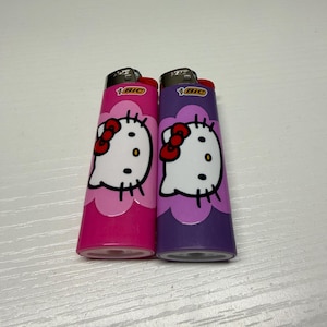 以下が含まれることがあります： ピンクと紫の2つのBICライター。それぞれにハローキティのデザインが施されています。ライターには赤いリボンと白い顔があります。ライターは白い表面にあります。