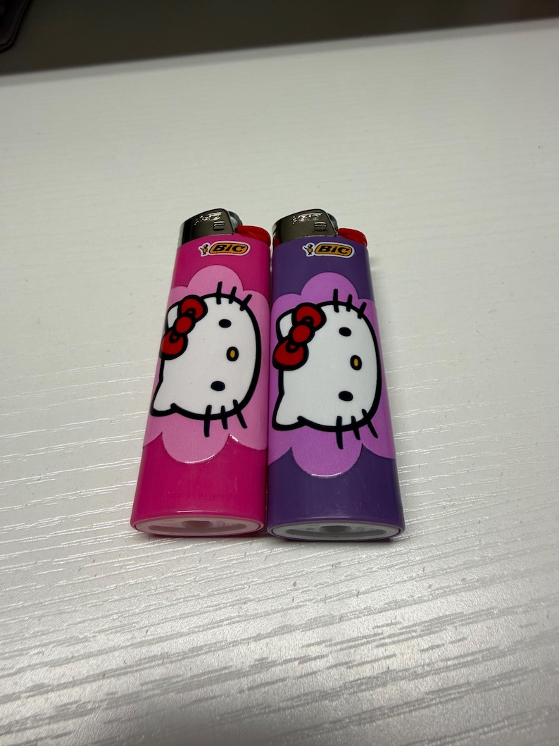 以下が含まれることがあります： ピンクと紫の2つのBICライター。それぞれにハローキティのデザインが施されています。ライターには赤いリボンと白い顔があります。ライターは白い表面にあります。
