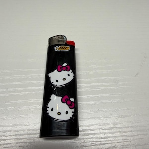 以下が含まれることがあります： ピンクのリボンが付いたハローキティの顔が2つ描かれた黒いBICライター。ライターには赤い点火ボタンとBICのロゴが付いています。