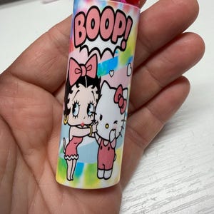 May include: A BIC lighter featuring a colorful tie-dye background with the text BOOP! in pink letters. The design includes a graphic illustration of Betty Boop and Hello Kitty standing together, both wearing pink bows, against a pastel-colored backdrop.