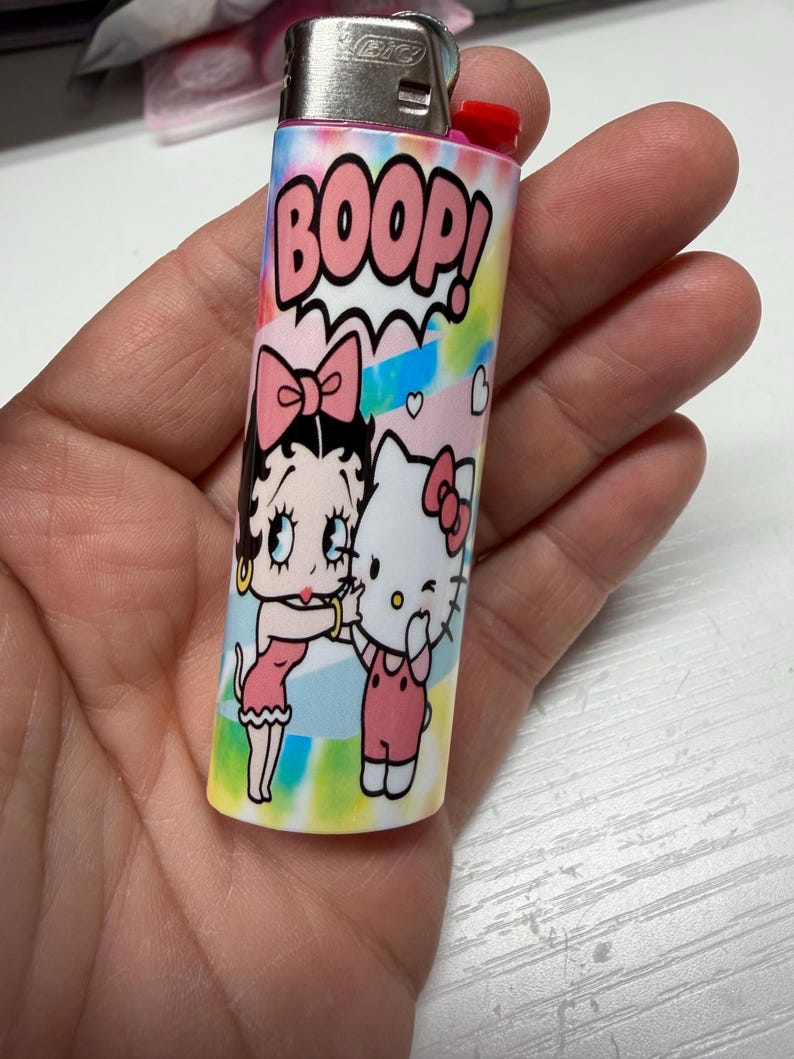 May include: A BIC lighter featuring a colorful tie-dye background with the text BOOP! in pink letters. The design includes a graphic illustration of Betty Boop and Hello Kitty standing together, both wearing pink bows, against a pastel-colored backdrop.