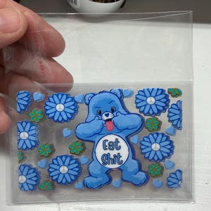 Op de afbeelding: Een doorzichtige plastic hoes met een verzameling blauwe beer-vormige stickers met de tekst Eat Shit op de buik, omringd door blauwe bloemstickers met vredestekens in het midden en kleine groene bloem- en blauwe hartvormige stickers.