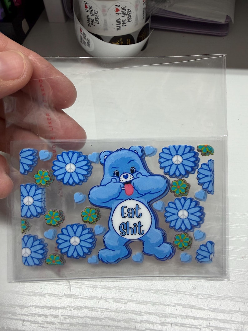 Op de afbeelding: Een doorzichtige plastic hoes met een verzameling blauwe beer-vormige stickers met de tekst Eat Shit op de buik, omringd door blauwe bloemstickers met vredestekens in het midden en kleine groene bloem- en blauwe hartvormige stickers.