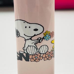 May include: A pink BIC lighter featuring a Snoopy graphic holding flowers. The design includes Snoopy, flowers, and the letter "N". The lighter has a red ignition button and a metal top.