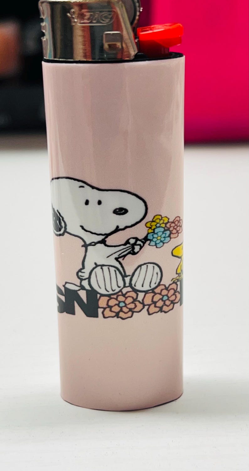 May include: A pink BIC lighter featuring a Snoopy graphic holding flowers. The design includes Snoopy, flowers, and the letter "N". The lighter has a red ignition button and a metal top.