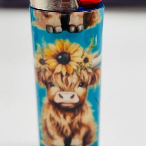 Pode incluir: Um isqueiro Bic azul com um design de vaca Highland adornado com uma coroa de girass&oacute;is. O isqueiro tem um bot&atilde;o de igni&ccedil;&atilde;o vermelho e uma parte superior de metal prateado. O desenho da vaca est&aacute; sobre um fundo turquesa.