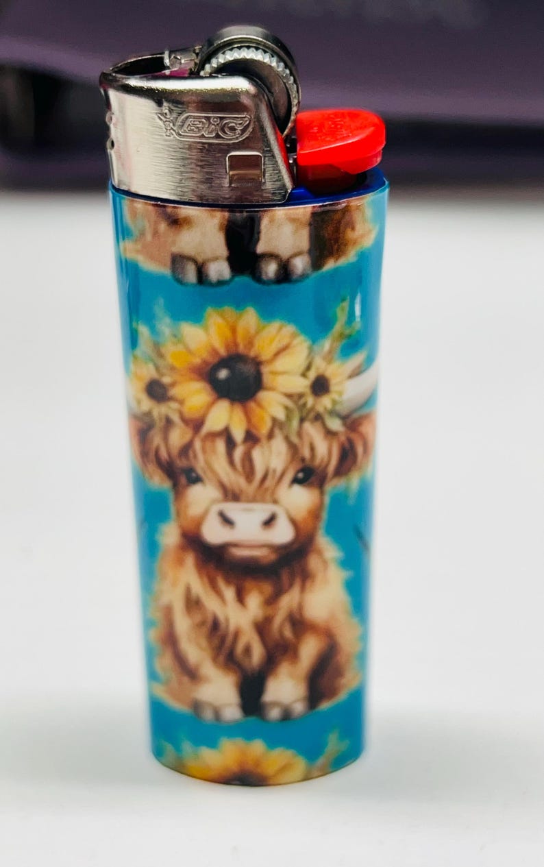 Pode incluir: Um isqueiro Bic azul com um design de vaca Highland adornado com uma coroa de girass&oacute;is. O isqueiro tem um bot&atilde;o de igni&ccedil;&atilde;o vermelho e uma parte superior de metal prateado. O desenho da vaca est&aacute; sobre um fundo turquesa.