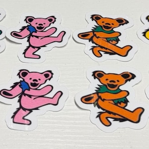May include: A collection of twelve stickers arranged in two rows. The set includes two circular stickers featuring a skull with a lightning bolt design, and ten die-cut stickers depicting dancing bears in green, pink, orange, yellow, and blue.