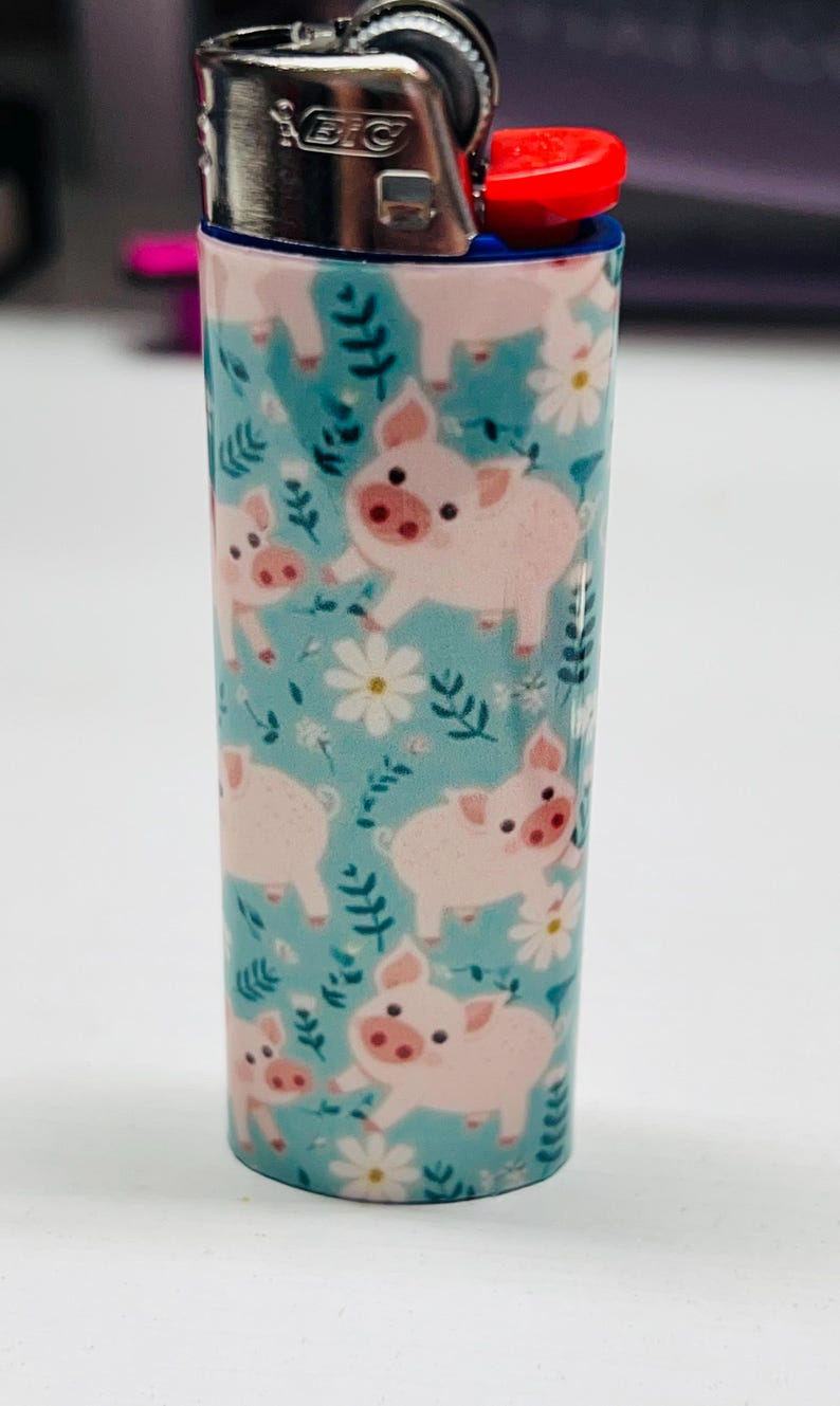 K&ouml;nnte beinhalten: Ein BIC-Feuerzeug mit einem skurrilen Design mit rosa Schweinen auf hellblauem Hintergrund. Das Feuerzeug hat einen roten Z&uuml;ndknopf und eine silberne Metalloberseite. Die Schweine-Illustration wiederholt sich auf der Oberfl&auml;che des Feuerzeugs.
