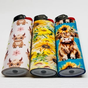 Pode incluir: Tr&ecirc;s isqueiros decorativos com designs &uacute;nicos. Um apresenta vacas das Terras Altas de desenho animado e flores cor-de-rosa sobre um fundo branco. Outro tem girass&oacute;is amarelos, e o terceiro uma vaca das Terras Altas com uma coroa de girass&oacute;is sobre um fundo azul.