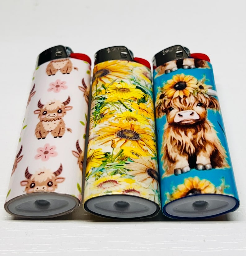 Pode incluir: Tr&ecirc;s isqueiros decorativos com designs &uacute;nicos. Um apresenta vacas das Terras Altas de desenho animado e flores cor-de-rosa sobre um fundo branco. Outro tem girass&oacute;is amarelos, e o terceiro uma vaca das Terras Altas com uma coroa de girass&oacute;is sobre um fundo azul.