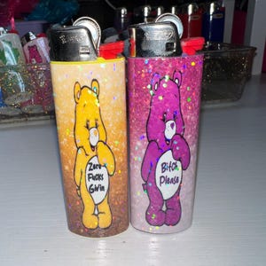 Puede incluir: Dos encendedores cubiertos de purpurina. Uno es amarillo con un oso de dibujos animados y el texto "Zero Fucks Givin". El otro es rosa con un oso morado y el texto "Bitch Please". Ambos tienen tapas de metal plateado.