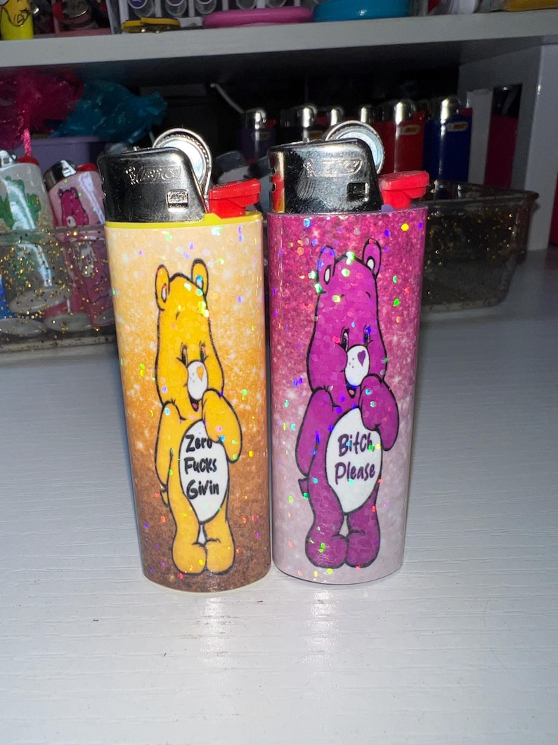 Puede incluir: Dos encendedores cubiertos de purpurina. Uno es amarillo con un oso de dibujos animados y el texto "Zero Fucks Givin". El otro es rosa con un oso morado y el texto "Bitch Please". Ambos tienen tapas de metal plateado.