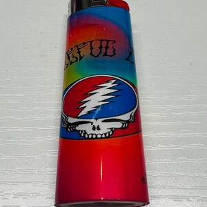 Puede incluir: Un encendedor colorido con un degradado rojo, azul y amarillo. El encendedor presenta un logotipo de calavera Steal Your Face en rojo, blanco y azul. El texto "STEAL" es visible.