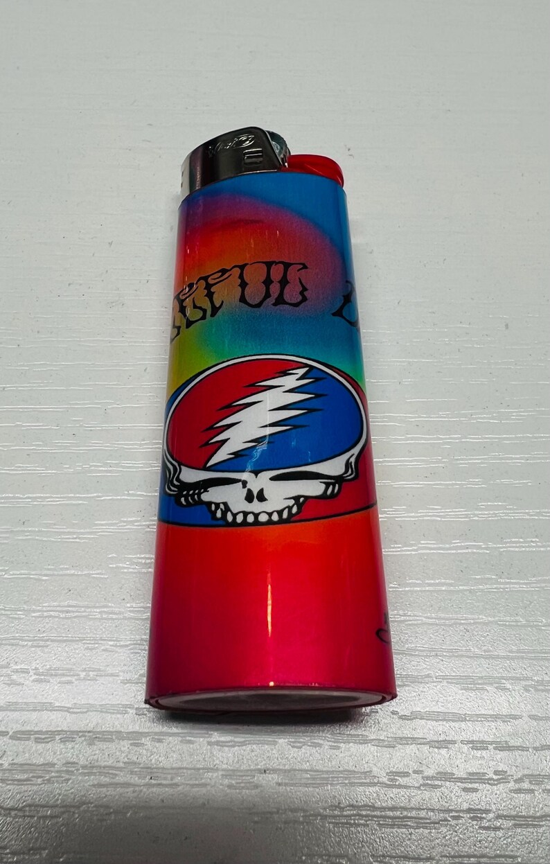 Puede incluir: Un encendedor colorido con un degradado rojo, azul y amarillo. El encendedor presenta un logotipo de calavera Steal Your Face en rojo, blanco y azul. El texto "STEAL" es visible.