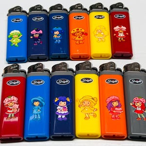 May include: A collection of colorful Scripto lighters featuring cartoon characters. The lighters are in various colors, including blue, red, orange, yellow, and gray. Each lighter has a different character design, with a silver metal top.