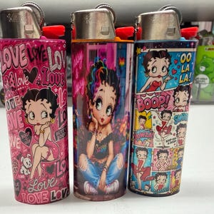 May include: Three Bic lighters featuring Betty Boop graphics. The left lighter is pink with the word LOVE and hearts. The center lighter shows Betty Boop in jeans and a black top. The right lighter features a comic strip design with the text OO LA LA.