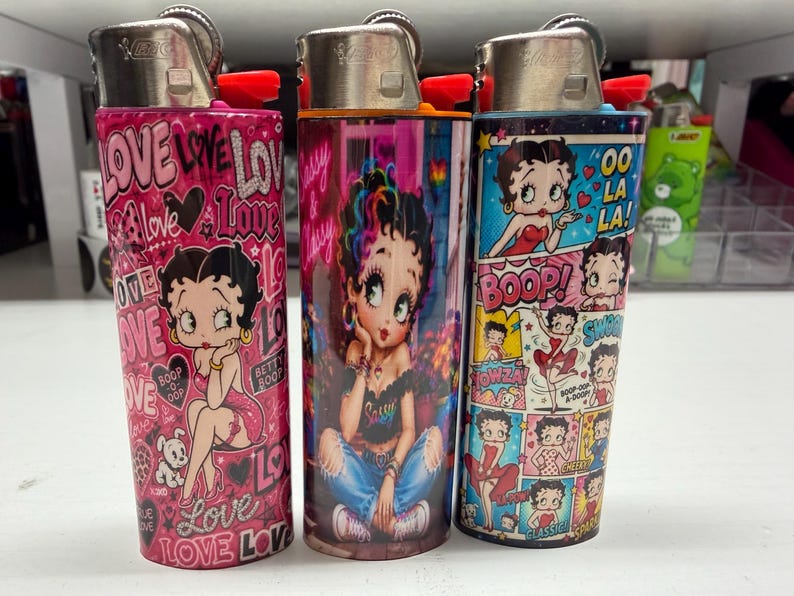 May include: Three Bic lighters featuring Betty Boop graphics. The left lighter is pink with the word LOVE and hearts. The center lighter shows Betty Boop in jeans and a black top. The right lighter features a comic strip design with the text OO LA LA.