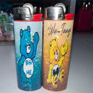 Puede incluir: Dos encendedores novedosos con dise&ntilde;os de osos de dibujos animados. Uno es azul con el texto "Bite ME", el otro es amarillo con el texto "Wu-Tang". Ambos encendedores tienen tapas plateadas y detalles rojos.