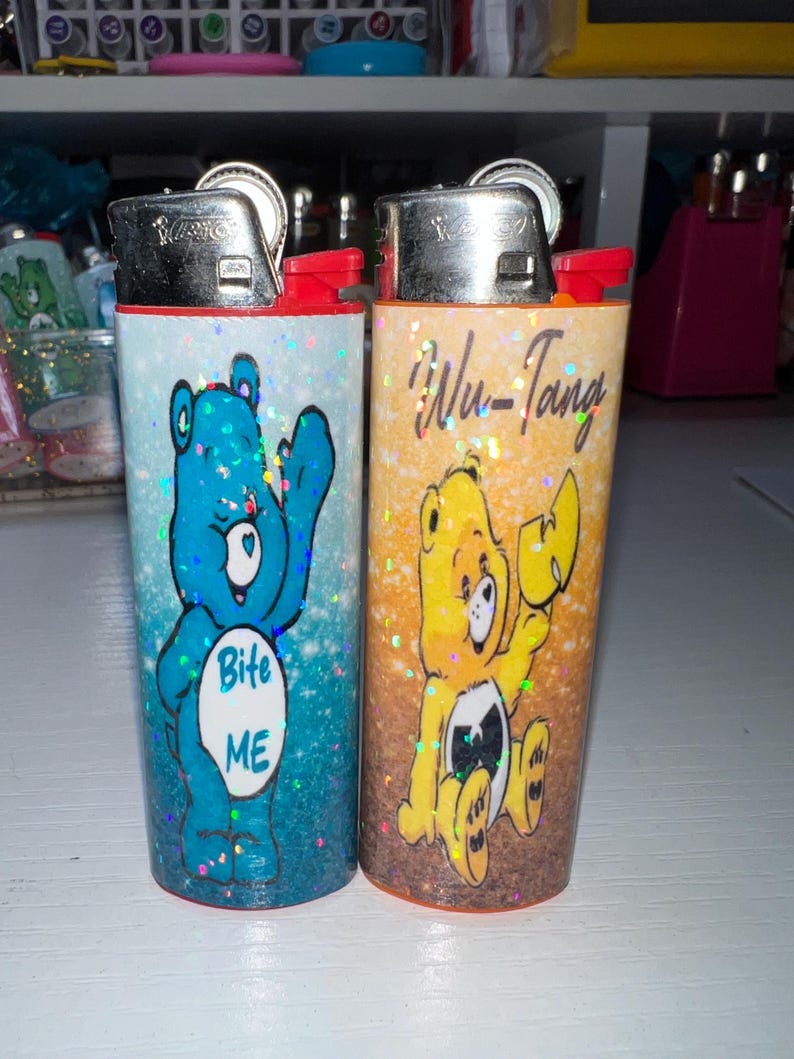 Puede incluir: Dos encendedores novedosos con dise&ntilde;os de osos de dibujos animados. Uno es azul con el texto "Bite ME", el otro es amarillo con el texto "Wu-Tang". Ambos encendedores tienen tapas plateadas y detalles rojos.