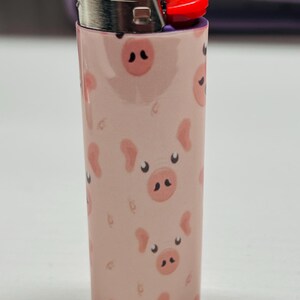 K&ouml;nnte beinhalten: Ein rosa BIC-Feuerzeug mit einem sich wiederholenden Muster aus Cartoon-Schweinegesichtern. Das Feuerzeug hat einen silbernen Metallaufsatz und einen roten Z&uuml;ndknopf. Die Schweinegesichter haben schwarze Nasenl&ouml;cher und kleine, gebogene Ohren.
